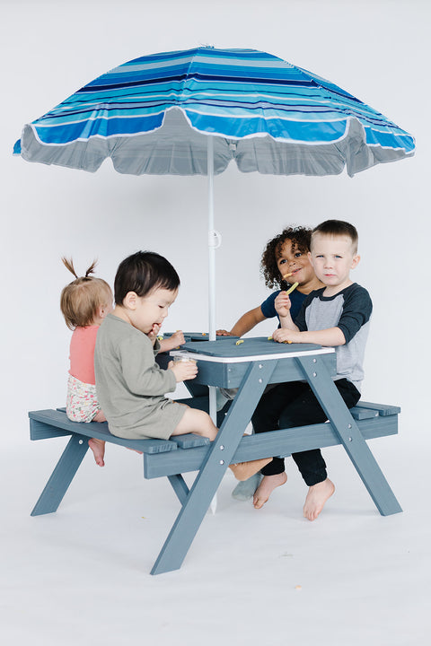 Four children gather around the Kids Picnic Table with 3 Collapsible Bins, engrossed in their activities. The compact blue table includes an adjustable umbrella adorned with blue stripes, providing shade and adding charm to their playful setting.