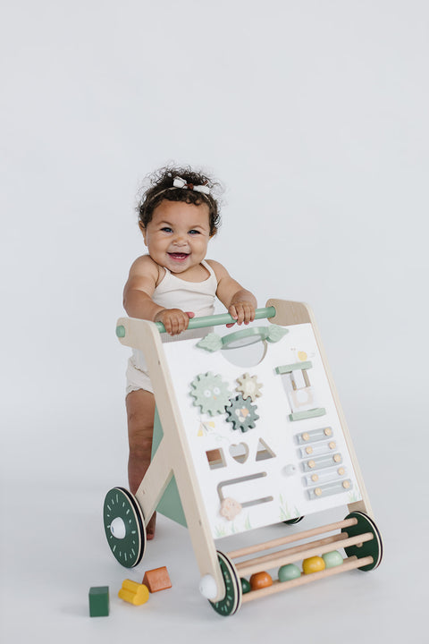 A baby stands smiling behind the high-quality "Wooden Baby Walker with Roller, Green," surrounded by scattered colorful blocks on a white background, promoting essential baby development skills with its various built-in toys.