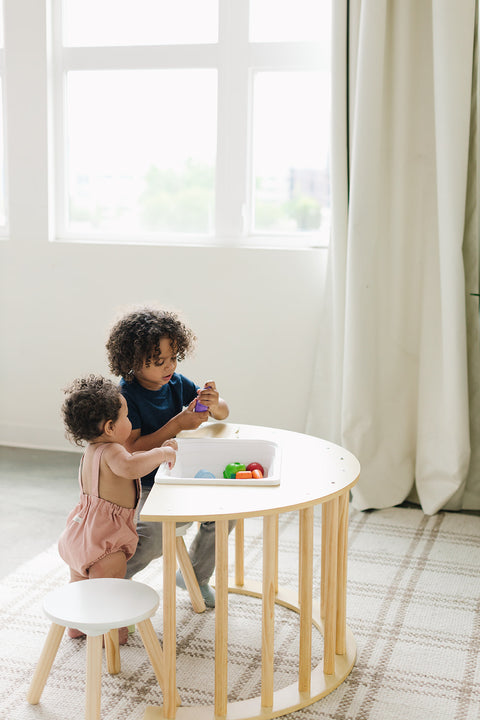 Two young children play together at a Climbing Arch Sensory Table with a bin, surrounded by natural light. Next to them, the table not only serves as a playful surface but also introduces an adventurous element that fosters creativity and balance, aligning with Montessori principles.