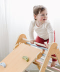 A toddler in a white shirt and pink pants climbs on the 5 In 1 Pikler Triangle Gym Set, which is part of a Montessori indoor playground.
