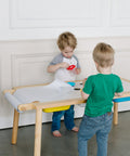 Two young children stand at the Painting Art Sensory Table with Paper Roll & Paint Cups + Bins, actively participating in a craft activity.