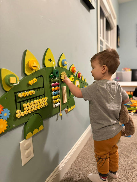 A child engages with the Dinosaur Activity Wall Busy Board Panels, skillfully manipulating gears and beads while clutching a stuffed animal in a room full of toys to enhance motor skills.