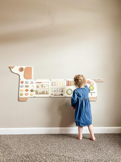 A child in a blue dress investigates the Dog Activity Wall Busy Board Panel, engaging with the sensory wall toy in a carpeted room with a beige backdrop.